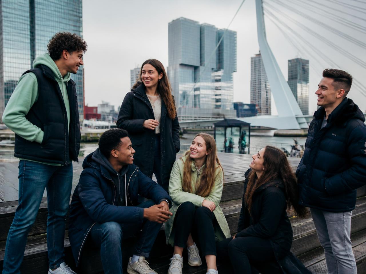 Groep jongeren in gesprek aan de Rotterdamse kade met de Erasmusbrug op de achtergrond